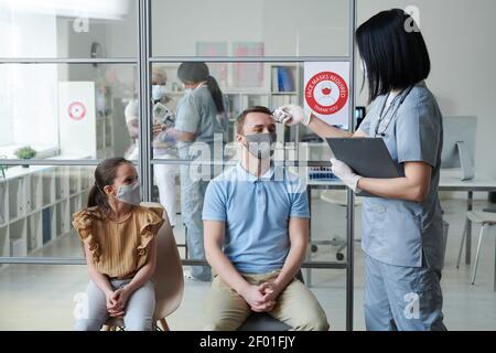 Jeune femme médecin ou infirmière en mesure uniforme de la température corporelle de l'homme dans le masque de protection et les vêtements décontractés assis à côté de sa fille Banque D'Images