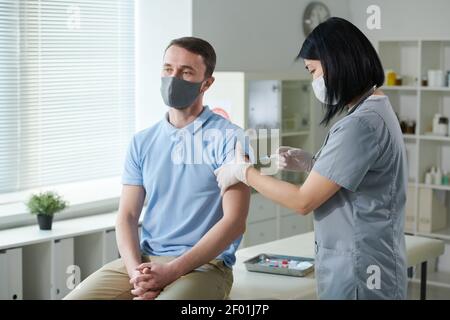 Brunette infirmière d'origine asiatique en uniforme et masque de protection vacciner un jeune homme assis dans le bureau médical des cliniques modernes Banque D'Images