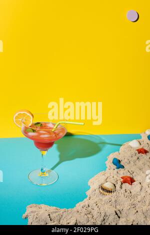 Cocktail rouge dans un grand verre avec une tranche d'orange à côté du sable et des coquillages sur une surface bleue sur un fond jaune avec un soleil violet. Créatif Banque D'Images