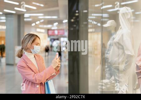 Jeune femme en masque faisant la photo de la nouvelle collection derrière la fenêtre pendant les achats dans le magasin Banque D'Images