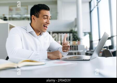 Un homme d'affaires confiant et satisfait lors d'un briefing vidéo. Un homme réussi s'assoit au bureau, communique avec ses collègues par conférence en ligne, montre ses pouces et ses sourires Banque D'Images