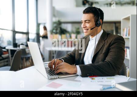 Homme d'affaires amical ou indépendant dans le casque, communique par appel vidéo. Un Manager ou un consultant professionnel séduisant s'assoit au bureau moderne avec un ordinateur portable, discutant en ligne Banque D'Images