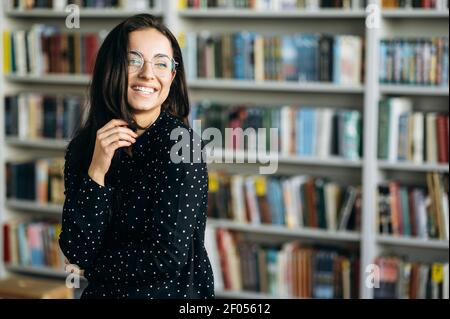 Joyeuse et élégante femme caucasienne indépendante ou étudiante, en lunettes et tenue formelle debout dans un bureau moderne ou une bibliothèque universitaire regarde et sourit Banque D'Images