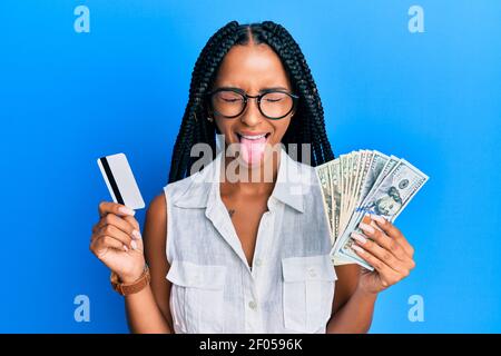 Belle femme hispanique tenant des dollars et carte de crédit collant la langue dehors heureux avec l'expression drôle. Banque D'Images