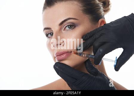 Jeune femme attrayante regarde la caméra tout en recevant une injection sur les lèvres par l'esthéticienne dans des gants médicaux de protection noirs. Lèvres augmentation conc Banque D'Images