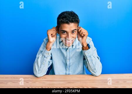 Jeune homme afro-américain élégant portant des vêtements décontractés assis sur la table souriant tirant les oreilles avec les doigts, geste drôle. Problème d'audition Banque D'Images