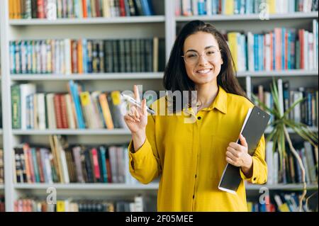 Portrait d'une magnifique jolie femme, debout dans un bureau ou une bibliothèque moderne. Jeune femme caucasienne en lunettes regarde la caméra, souriant. Enseignante ou pigiste Banque D'Images