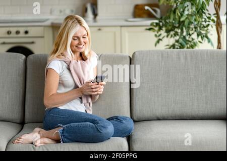 La femme blanche, blonde et mûre, est assise sur le canapé, à l'aide d'un smartphone. Bonne femme d'âge moyen chat avec des amis ou la famille, la navigation sur Internet, sourire Banque D'Images