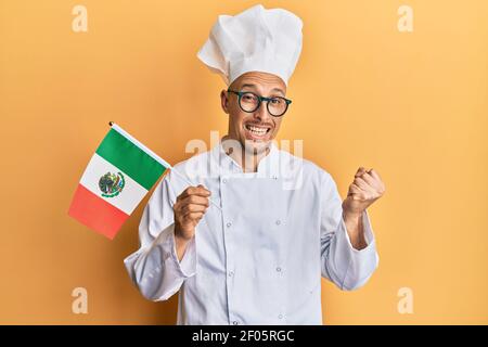 Homme chauve avec barbe portant un tablier de cuisinier professionnel tenant le drapeau du mexique criant fier, célébrant la victoire et le succès très excité avec le bras levé Banque D'Images