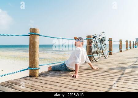 Portrait de l'homme relaxant habillé de vêtements d'été légers et lunettes de soleil assis et profiter du temps et de la vue sur la plage sur le bois pier.négligent vacances à t Banque D'Images