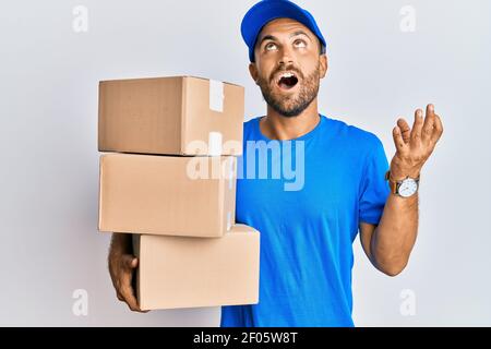 Beau homme avec la barbe portant l'uniforme de messagerie tenant les colis de livraison hurlant et hurlant, fou et fou, avec une expression agressive et les bras se relèvent Banque D'Images