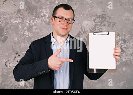 Jeune homme d'affaires ou gérant joyeux en lunettes et costume, tenant le presse-papiers avec une feuille de papier propre et pointant son doigt contre lui debout Banque D'Images