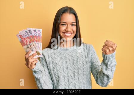 Belle femme hispanique tenant 100 billets de hong kong dollars criant fier, célébrant la victoire et le succès très excité avec le bras relevé Banque D'Images