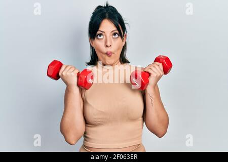 Jeune femme hispanique portant des vêtements de sport en utilisant des haltères faisant le visage de poisson avec la bouche et les yeux de bourdonner, fou et comique. Banque D'Images