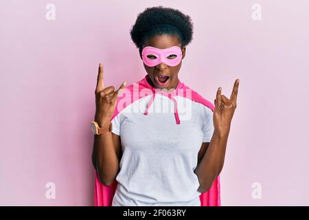 Jeune fille afro-américaine portant un masque super-héros et un déguisement de cape criant avec une expression folle faisant le symbole du rock avec les mains vers le haut. Star de la musique. Heavy Banque D'Images