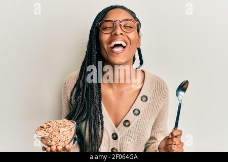 Femme afro-américaine avec des tresses mangeant des céréales complètes saines souriant et riant fort parce que drôle de blague folle. Banque D'Images