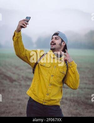 Jeune homme souriant routard dans une veste en Jean jaune prenant le selfie sur un smartphone en se tenant sur un pré brumeux dans la nature Banque D'Images
