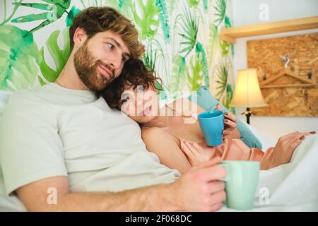 Jeune couple en vêtements de nuit enserrer tout en appréciant le café chaud à l'intérieur lit sur draps blancs avec oreillers et vue sur la caméra dans l'appartement de lumière Banque D'Images