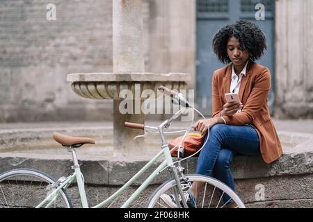 Jeune femme afro-américaine dans une tenue élégante et décontractée naviguant sur mobile téléphone tout en étant assis près d'un vélo sur la vieille bordure de fontaine en pierre en ville Banque D'Images