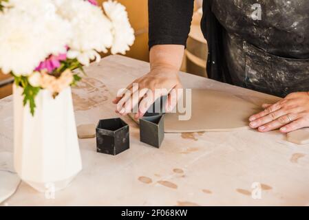 Cachez une femme non reconnaissable avec un tablier et une tenue décontractée noire objets géométriques pour couper des formes dans l'argile sur la table près de vase avec fleurs en travail Banque D'Images