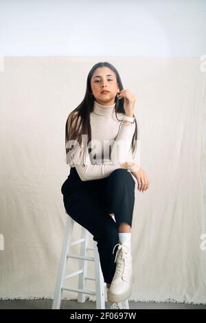 Jeune femme peu émotive et réfléchie regardant un appareil photo assis sur un tabouret en arrière-plan studio en journée Banque D'Images