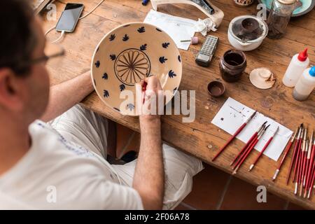 Depuis le dessus de la récolte, homme sans visage assis à la table avec pinceaux et croquis dessinés sur une plaque en céramique faite à la main Banque D'Images