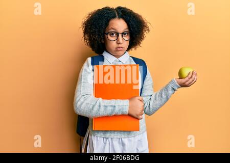 Jeune fille avec cheveux afro portant sac d'école tenant des livres et pomme verte dans le visage de choc, regardant sceptique et sarcastique, surpris par ouvert Banque D'Images