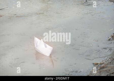 Le bateau en papier blanc fait main navigue jusqu'au rivage. Eau sale et floes de glace. Concept du début du printemps, liberté. Banque D'Images