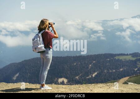 Jeune femme élégante et confiante avec un sac à dos dans les montagnes regarde le paysage à travers des jumelles. Voyage seul autour du quartier. Wanderl Banque D'Images