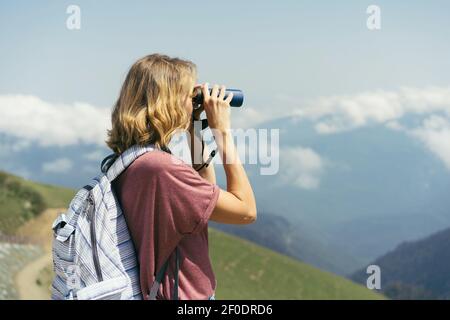 Une femme de randonnée regarde à travers des jumelles tout en se tenant debout dans les montagnes. Tourisme en montagne en été. Banque D'Images
