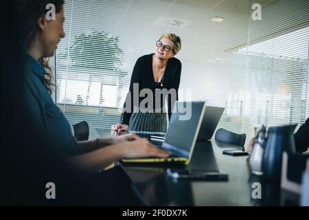 La haute direction se tient à la table de la salle de réunion et regarde sa collègue. Équipe de professionnels en voyage d'affaires ayant une réunion dans une salle de conférence de bureau. Banque D'Images