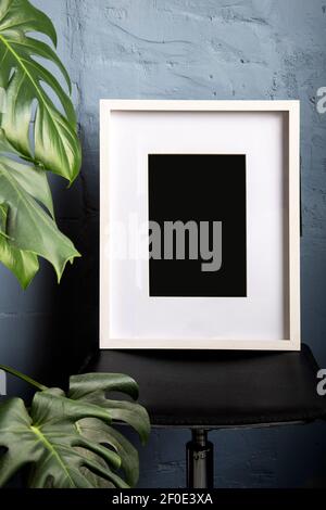 Maquette d'affiche intérieure avec cadre en bois blanc vide vertical sur chaise noire, fleur de monstère, plante devant dans la chambre avec mur bleu Banque D'Images