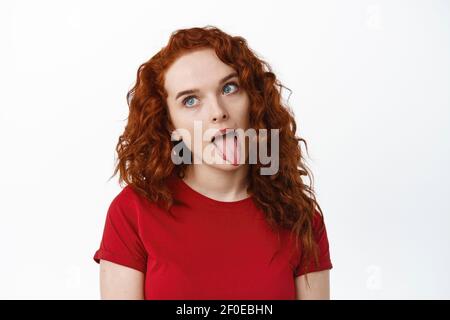 Portrait d'une fille de gingembre stupide montrant la langue et l'idiot autour, faisant des visages drôles et regardant de côté, debout sur fond blanc Banque D'Images