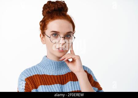 Portrait d'une jeune femme réfléchie à tête rouge dans des lunettes, ayant ses cheveux dans un petit pain, touchant le visage tout en pensant, regardant l'appareil photo déterminé, blanc Banque D'Images