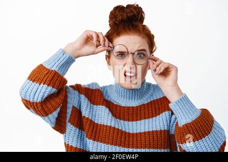 Jeune fille nerdy avec des cheveux de gingembre et des yeux bleus, essayant de porter des lunettes croquées, ne peut pas voir sans lunettes, debout drôle contre blanc Banque D'Images