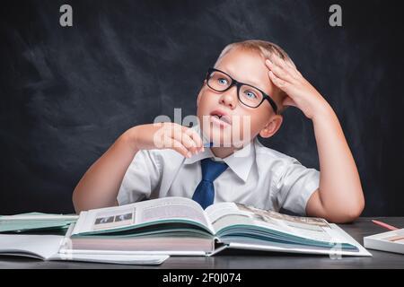 Un écolier, un caucasien dans une chemise blanche avec une cravate, assis à une table portant des lunettes, lit un livre. Un visage surpris et un look expressif. Noir Banque D'Images