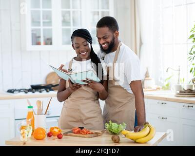 Joyeux couple noir lisant la recette dans le livre de cuisine tout en cuisinant le déjeuner dans la cuisine Banque D'Images