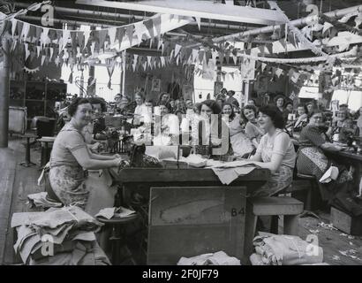 1953, image historique, souriant visages comme les travailleuses de la compagnie de drapery, Hepworths de Leeds, Angleterre, Royaume-Uni, assis à leurs bancs de travail avec des machines à semer. Le plafond de l'usine est décoré de banderoles et de drapeaux célébrant le couronnement de la reine Elizabeth au trône britannique qui tok place le 2nd juin 1953 à l'abbaye de Westminster. La Providence Works sur Clay Pit Lane était l'emplacement de l'entreprise de Joseph Hepworth et son, une compagnie de vêtements a commencé en 1865 et qui en 1905 avait 143 magasins. Plus tard dans le siècle, Hepworths est devenu Next. Banque D'Images