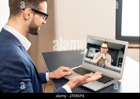 Employé réussi avec des lunettes en costume élégant, assis à l'ordinateur portable, réunion d'affaires en ligne avec des clients et partenaires potentiels, regardant l'ordinateur portable, travaillant à distance Banque D'Images