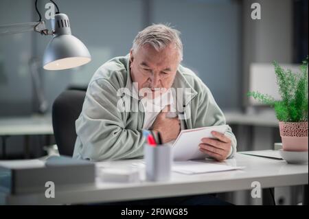Dans son bureau, un homme aux cheveux gris a ressenti une douleur dans son cœur en travaillant, il est assis avec sa main sur sa poitrine Banque D'Images