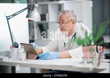 Un médecin âgé voit des patients en ligne à l'aide d'une tablette et d'un appel vidéo. Assistance à distance d'un spécialiste médical. Concept e-santé Banque D'Images