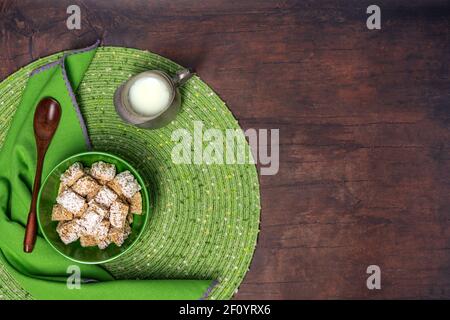 petit déjeuner de céréales de blé dépolies dans un bol vert avec un napperon, une serviette, une cuillère en bois et un petit pichet de lait en étain sur fond de table en bois Banque D'Images