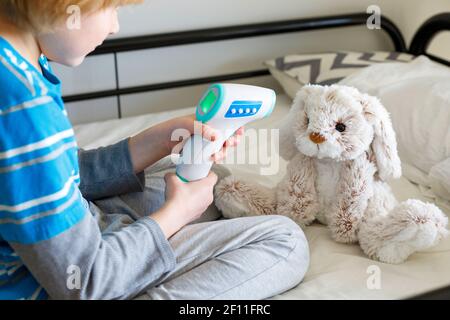 Le petit enfant mesure la température du lapin moelleux avec un thermomètre sans contact. Mignon petit enfant assis sur le lit, jouant docteur avec son jouet bourré Banque D'Images