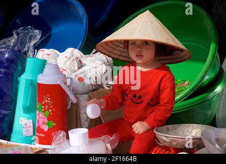 Jeune enfant vietnamien dans une cabine de marché portant des vêtements rouges et un chapeau conique de style vietnamien distinctif. Bac Ha, province Lao Cai, Vietnam Banque D'Images