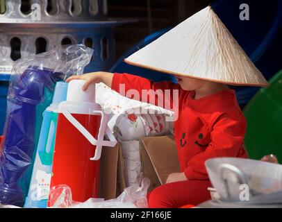 Jeune enfant vietnamien dans une cabine de marché portant des vêtements rouges et un chapeau conique de style vietnamien distinctif. Bac Ha, province Lao Cai, Vietnam Banque D'Images