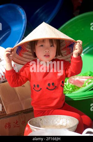 Jeune enfant vietnamien dans une cabine de marché portant des vêtements rouges et un chapeau conique de style vietnamien distinctif. Bac Ha, province Lao Cai, Vietnam Banque D'Images