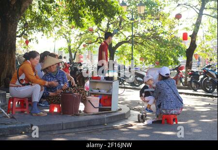 Vie de rue vietnamienne. Vendeurs vendant des collations sur le front de mer, Hoi an, Vietnam. Mère allaitant garçon en pram. Banque D'Images