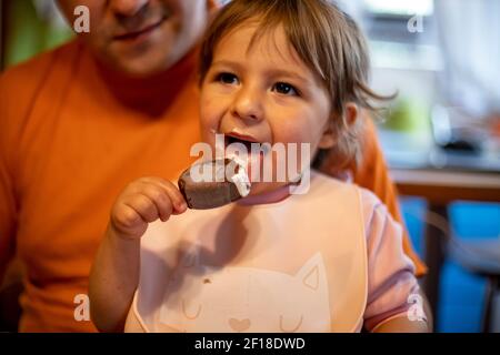 un adorable tout-petit aime manger de la glace à la popsicle sur les genoux de ses parents dans l'intérieur de la maison Banque D'Images