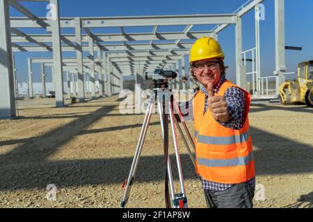 Employé dans un casque rigide jaune montrant une affiche de main à pouce vers le haut. Happy mature Land Surveyor avec tacheomètre regardant la caméra. Banque D'Images