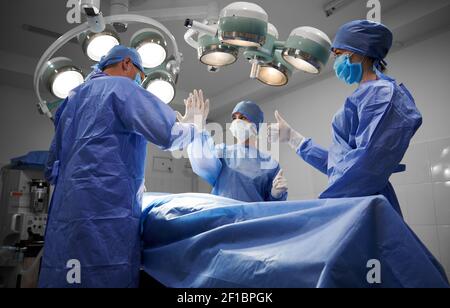 Médecins debout par le patient après une opération chirurgicale réussie. Chirurgien plastique donnant cinq hauts à l'assistant. Deuxième assistant faisant un geste du pouce vers le haut. Concept de médecine et travail d'équipe réussi. Banque D'Images